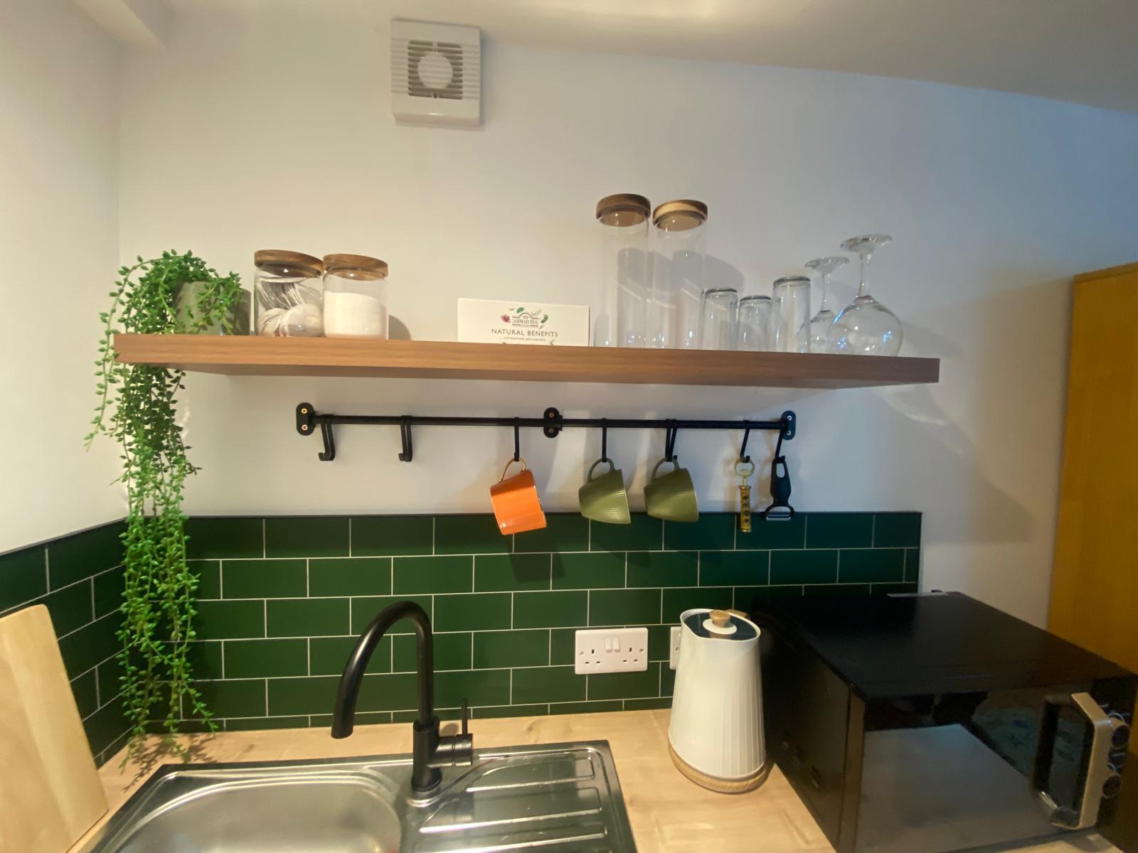 Kitchenette with green metro tiles, an oak shelf, a black tap and a stainless sink